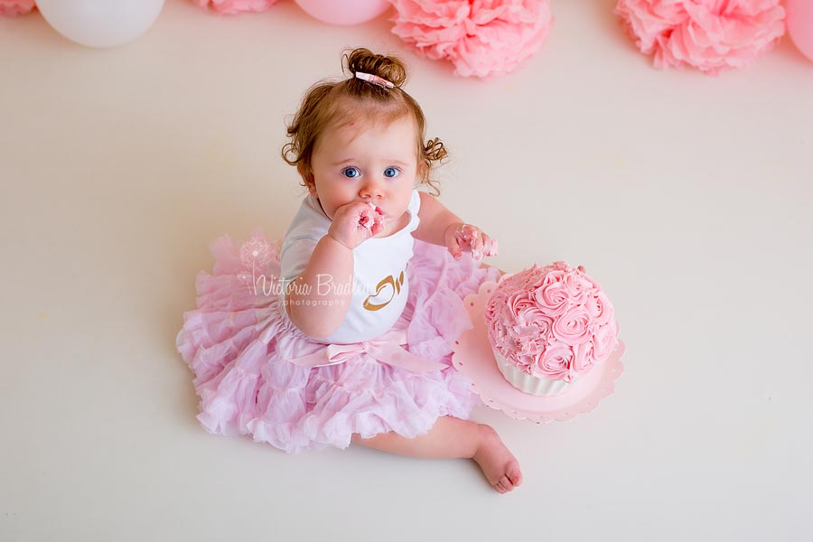 Baby sat on the floor in pink tutu and number one on her top, eating a pink and white cake