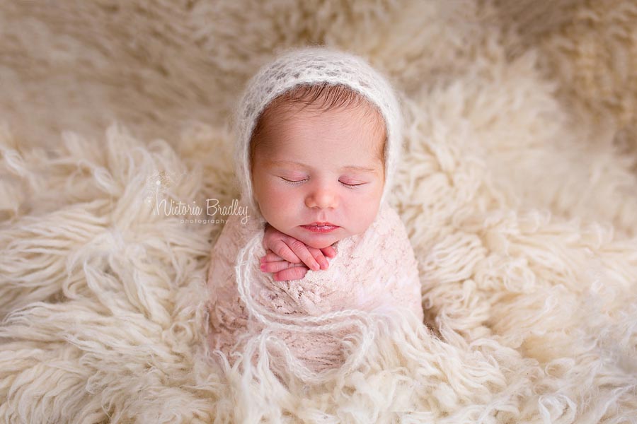 newborn baby girl in the potato sack pose on a cream flokati rug wearing a white mohair bonnet with a blush lace wrap