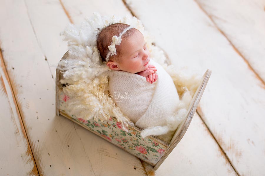 baby in a floral newborn photography crib prop, wrapped in a cream wrap with a bow detail tie back on a cram wood floor