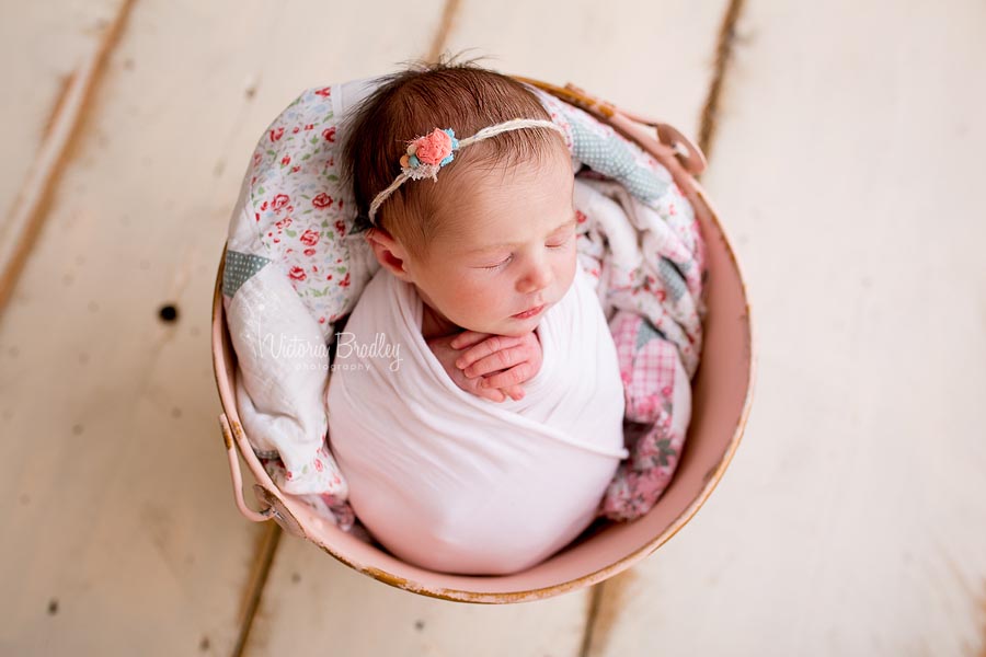 newborn baby in a vintage pink pail swaddled in a pink wrap placed on a wooden painted floor