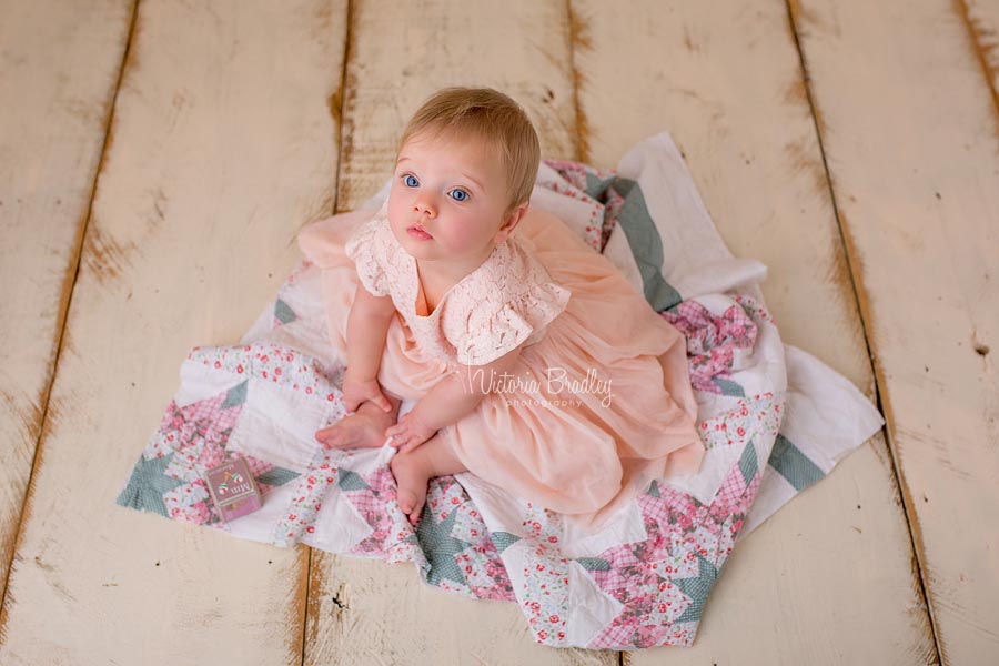 7 month baby girl sat on a pink and sage vintage quilt layer with a peach coloured dress on cream wooden floor boards