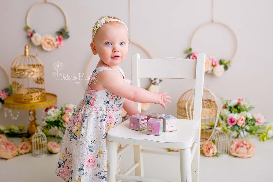 boho floral cake smash with 1 year old baby girl, with flowers, gold bird cages and floral hoops, stood by a white chair