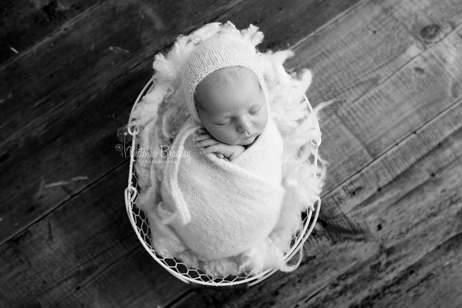 black and white image of a swaddled newborn boy in cream wrap & bonnet on a cream felted curls stuffer in a basket on dark wooden floor boards 