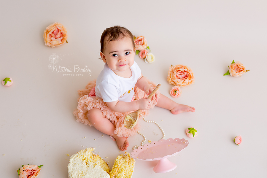 baby playing with wooden spoon and pearls, with a peach tutu and white top with a sponge cake and pink cake stand