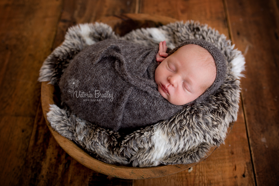 wrapped newborn baby boy on faux fur with dark grey knitted wrap and bonnet in a wooden bowl and a wooden floor