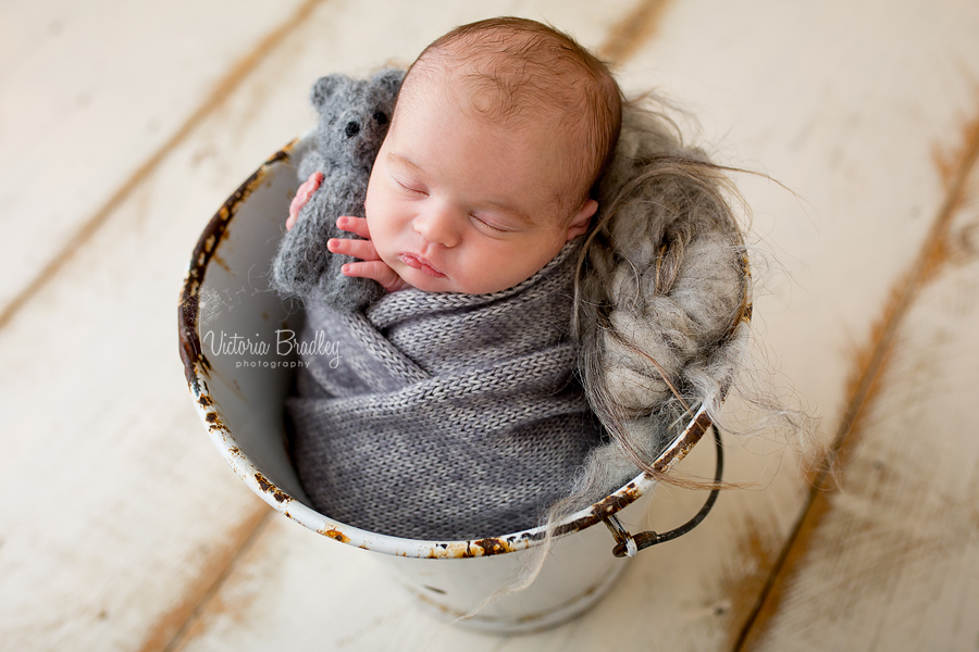 newborn baby boy in grey knitted wrap in a white vintage bucket holding a little knitted grey bear