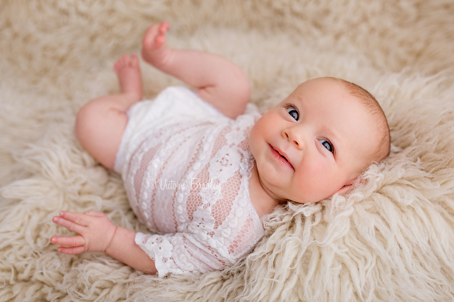 awake 7 week old baby smiling on cream flokati rug, wearing a lace romper