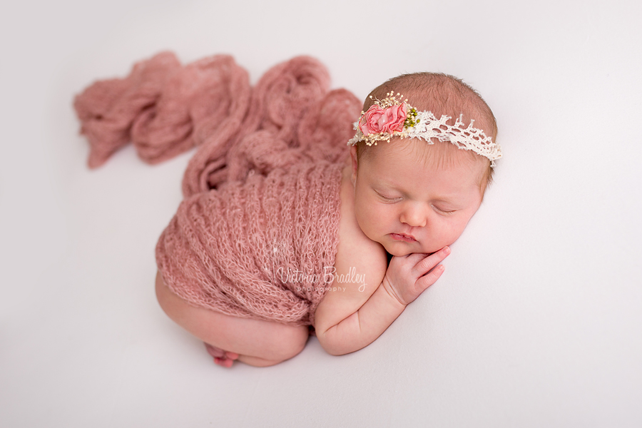 newborn baby lay on her tummy with a pink rose tieback, dusky pink wrap on a cream fabric backdrop