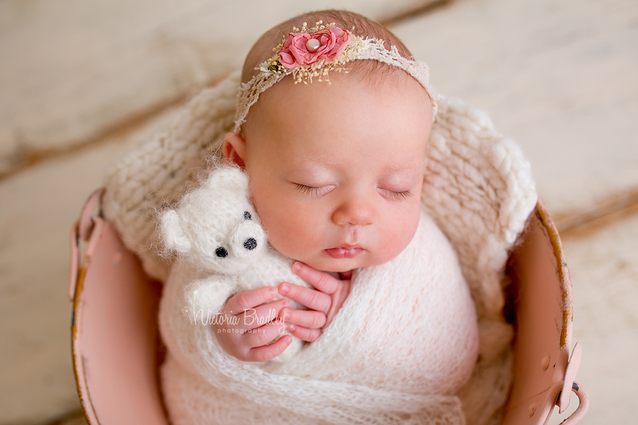 sleepy older newborn holding small cream teddy bear in pink vintage metal bucket