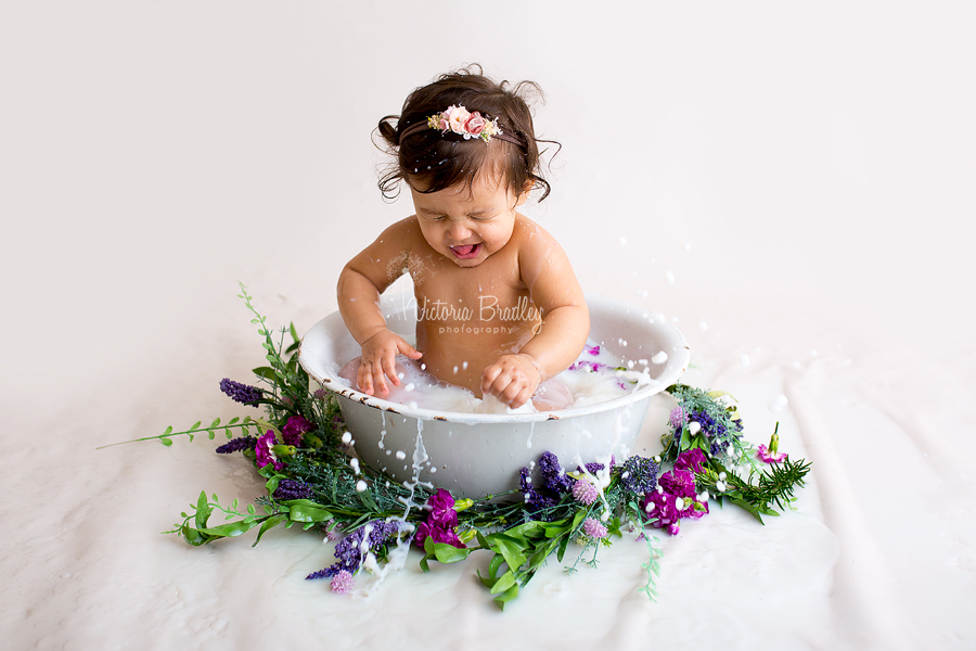 baby girl splashing during a milk bath photography session, using a white enamel bath tub with purple and green flowers and foliage 