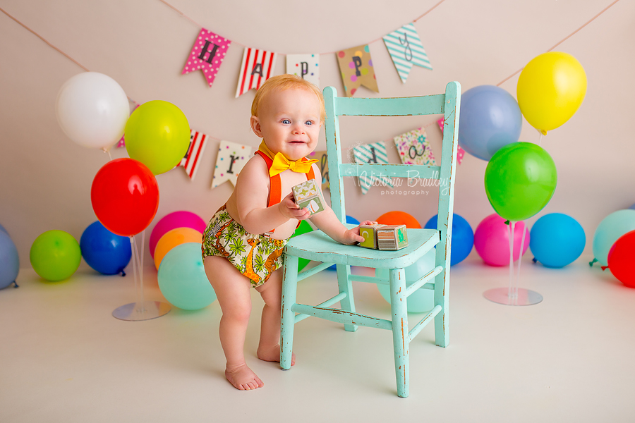 baby boy cake smash, stood next to an aqua chair, holding blocks