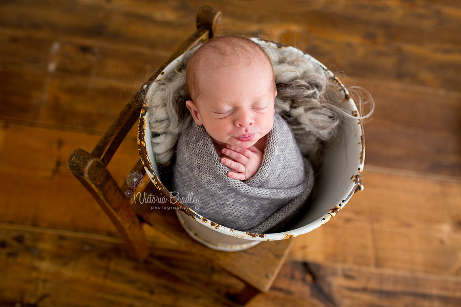 wrapped baby newborn in grey knitted wrap in a white bucket on a wooden chair