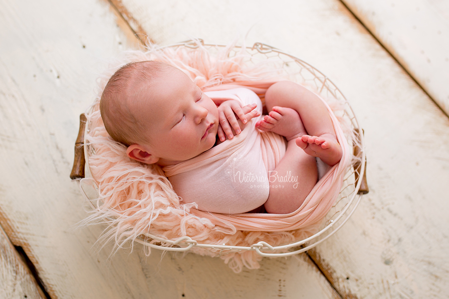 newborn girl in cream egg basket with peach fur stuffer on cream wood floor