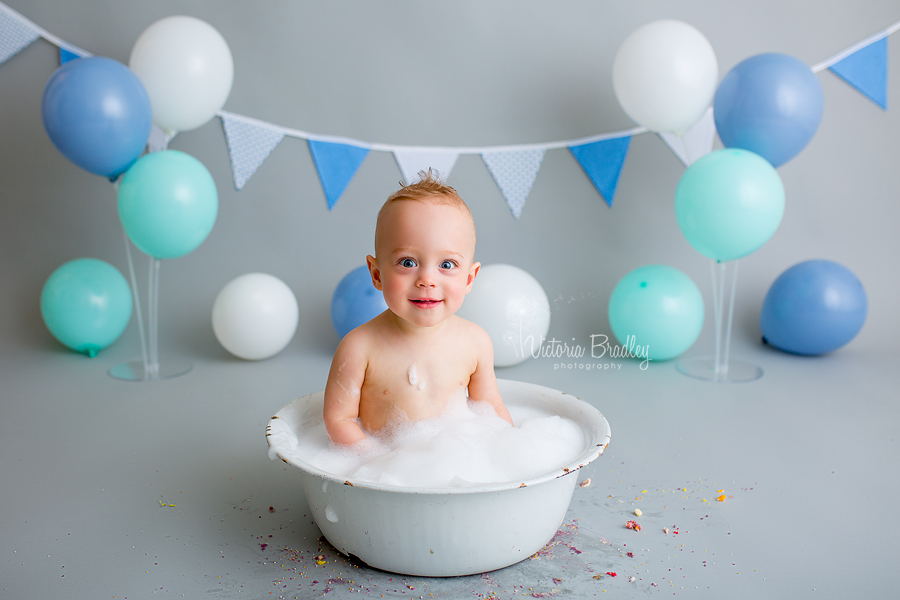 baby boy in vintage bath tub with bubbles on grey backdrop with balloons