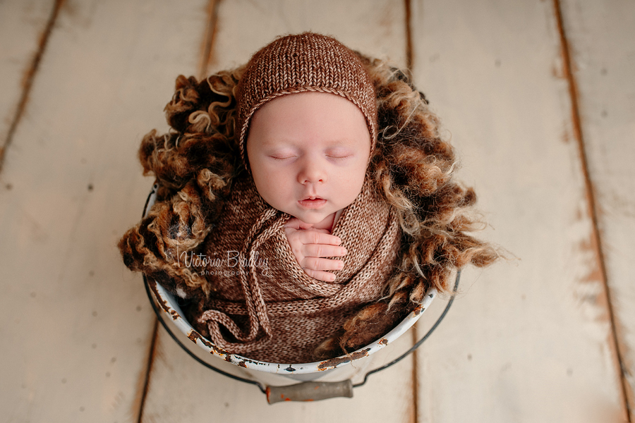 newborn baby boy in brown flecked wrapped and bonnet in white bucket with brown curly fur layer