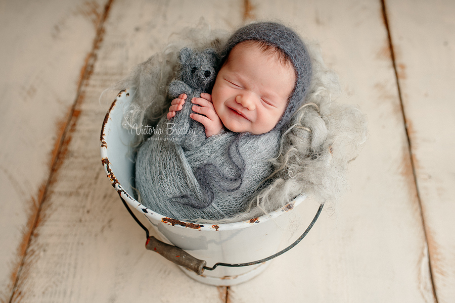 wrapped newborn baby girl newborn photography in grey wrap with grey teddy in white bucket on cream floor boards