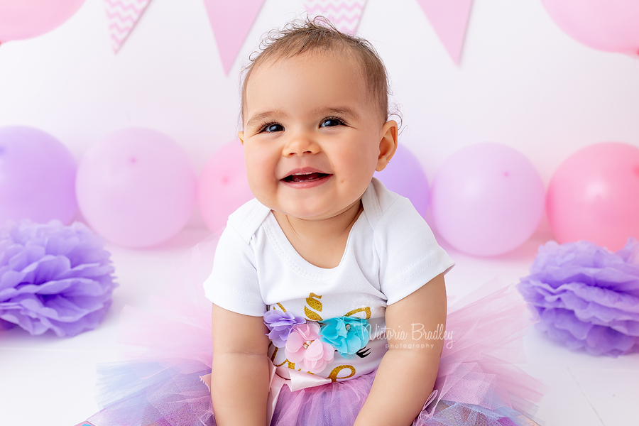 baby girl cake smash sat on floor with pink and purple ballooos