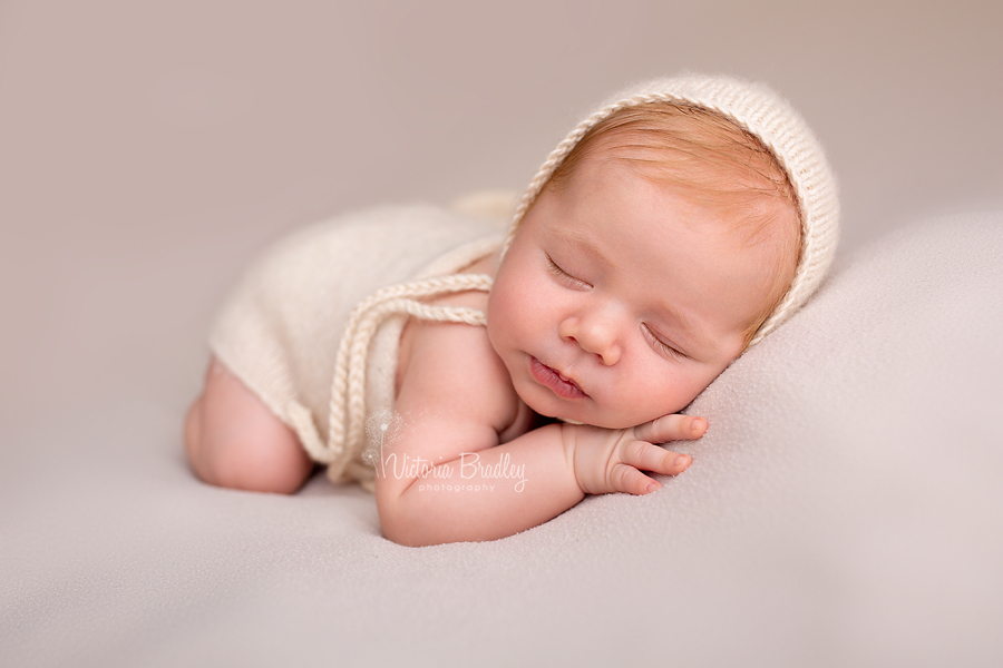 asleep newborn on tummy pose on cream backdrop with cream bonnet